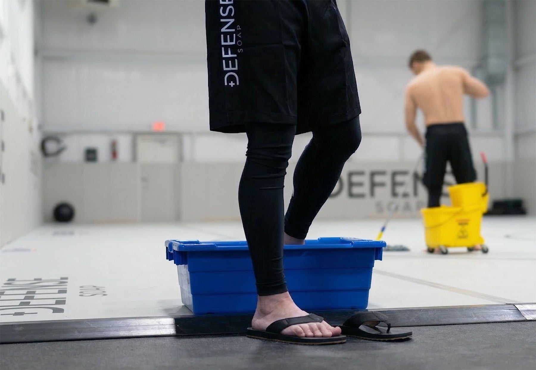 A Jiu-jitsu player using a Defense Foot Cleaning Station on light grey mats after rolling, while another person mops in the background of a professional combat sports facility.